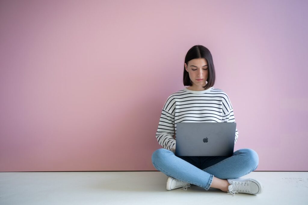 brunette woman working on laptop on floor, pink wall. how to earn 5,000 dollars per month