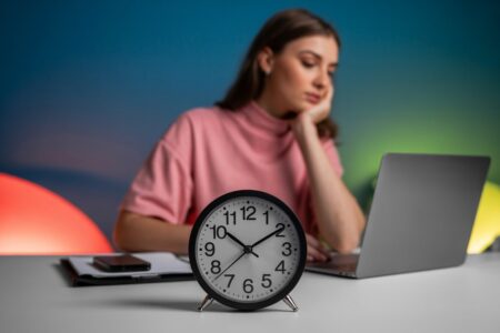 clock in the foreground, young woman in pink t-shirt working on laptop, neon background. how to make $5,000 fast
