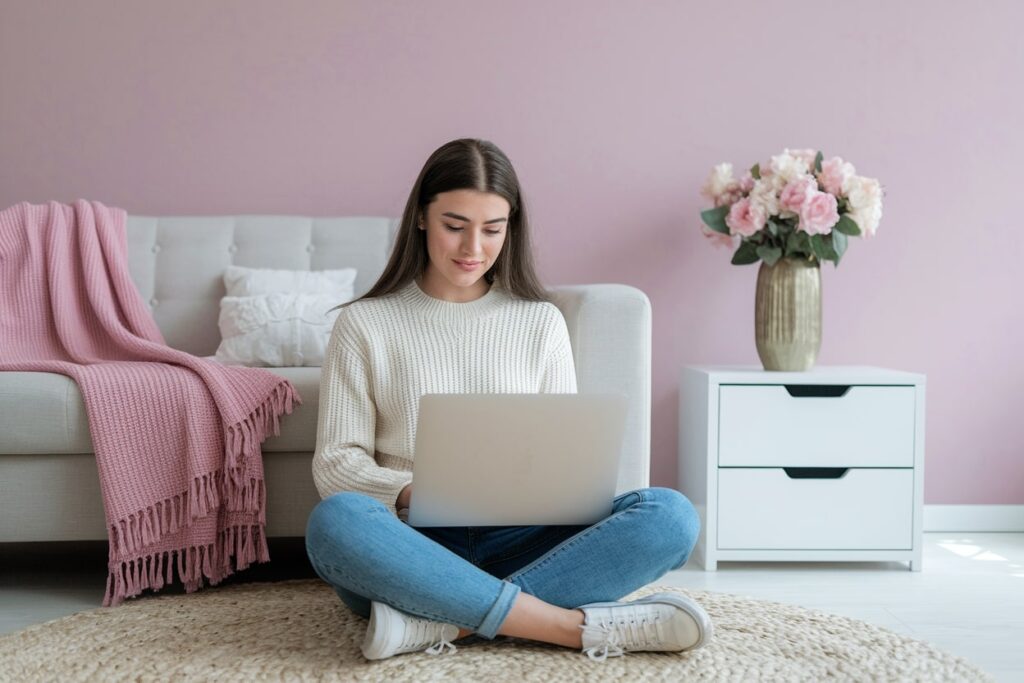 young female in light sweater and jeans sitting with laptop on the floor in the living room, pink wall. how to make $5k a month