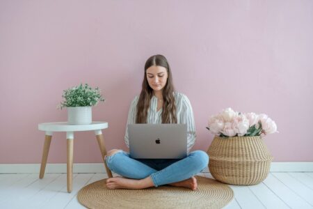 young female sitting with laptop on the floor, pink wall, white floor, flowers, side table. how to make $5,000 a month