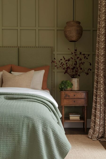 earthy bedroom has deep sage green wall with board and batten, terracotta and cream pillows, wood nightstand