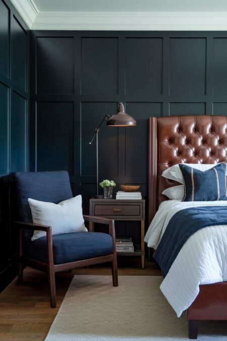 masculine bedroom with navy blue walls, tufted leather headboard, and wooden armchair with a dark blue cushion