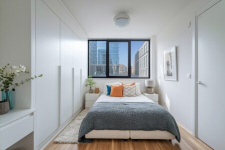 modern small bedroom with white built-in wardrobe, black window frame, gray blanket, and orange accent pillows