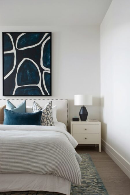 small bedroom with a large dark artwork above headboard, white walls, dark blue accent pillow, and black table lamp