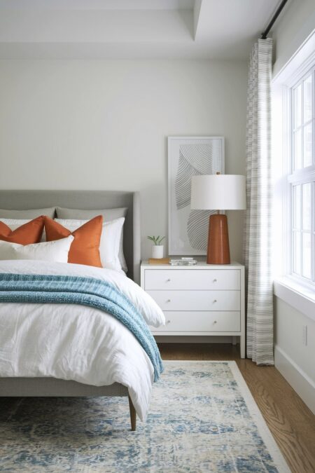 small bedroom with white walls, gray upholstered bed frame, burnt orange accent pillows and table lamp