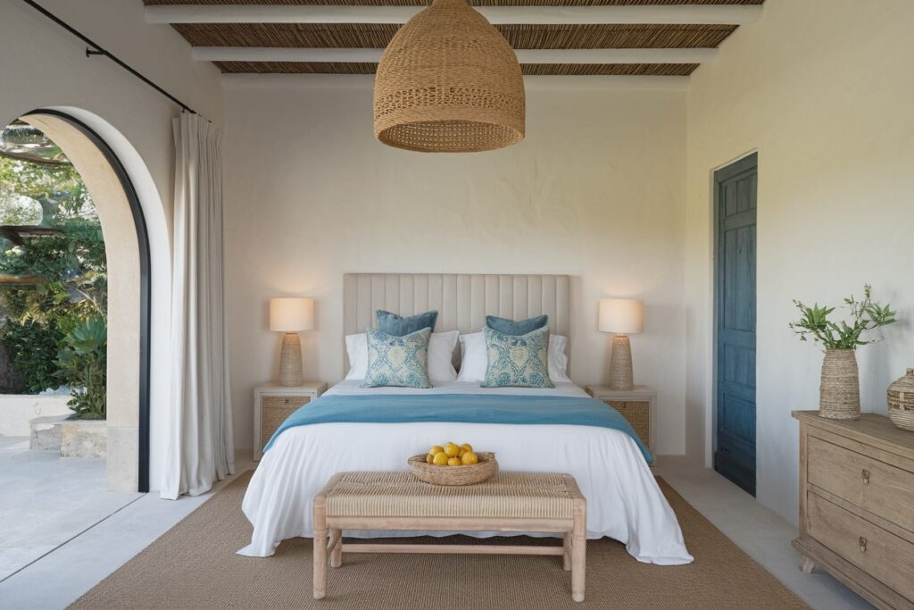 13 Mediterranean Bedroom Ideas for a Serene, Sun-Washed Space Mediterranean bedroom with white bedding, blue throw pillows and blanket, channel tufted headboard, and woven pendant light