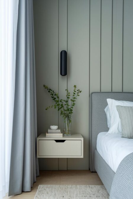 modern bedroom with sage green walls, white floating nightstand with a drawer, gray upholstered bed frame, and a black wall sconce