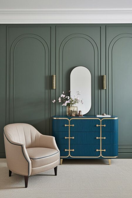 sage green wall with decorative arches, blue curved Art Deco dresser with gold hardware, and a cream armchair with a rounded back