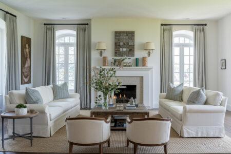 French country style living room with white walls, fireplace, cream slipcovered sofas with gray pillows, curved armchairs