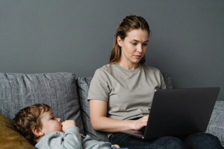 34 Side Hustles for Single Moms Who Want Extra Income mom works on laptop while sitting on a gray sofa at home