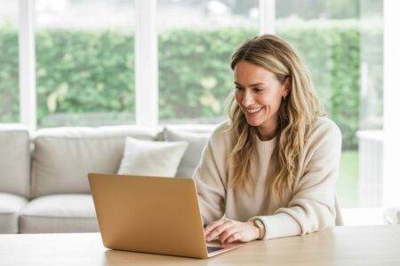 smiling blonde female in a light color sweater working on the laptop at home