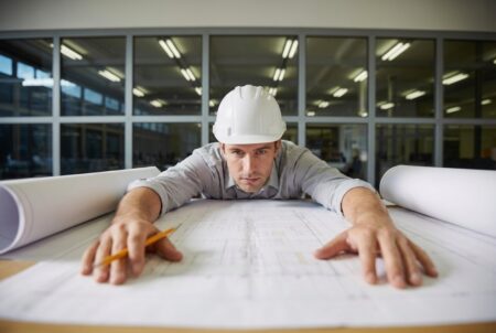 Side hustles for architects. A young architect in a white hard hat is looking at the blueprints.
