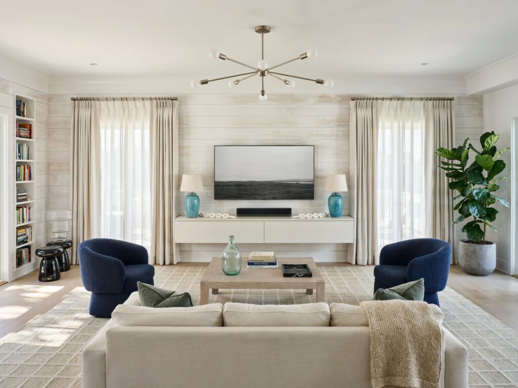 beach house living room with whitewashed shiplap walls, navy blue swivel barrel chairs, a TV, and floating media console with two table lamps with rounded ceramic bases in an aqua-blue crackle glaze