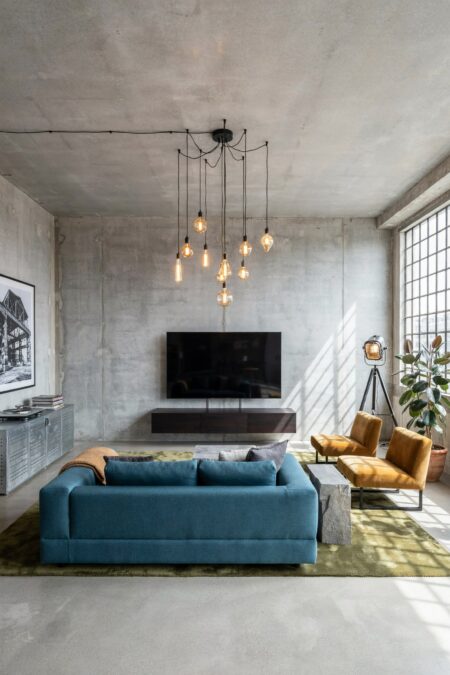 industrial living room in concrete, pendant lights with Edison bulbs, a teal-blue sofa, mustard yellow accent chairs, and a moss green rug
