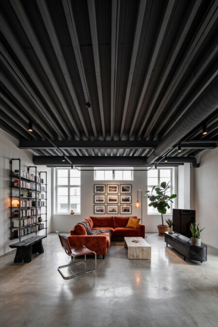 industrial living room with matte black ceiling beams and ductwork, a rust-orange sofa, a perforated media console, pipe shelving, and a black-and-white photography gallery wall