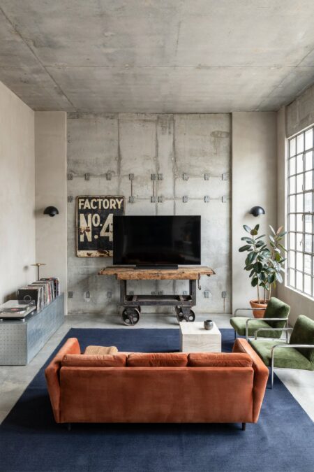 industrial living room with a rust-orange velvet sofa, a weathered factory cart media console, a vintage tin sign on the wall behind the TV, a navy blue rug, and green accent chairs