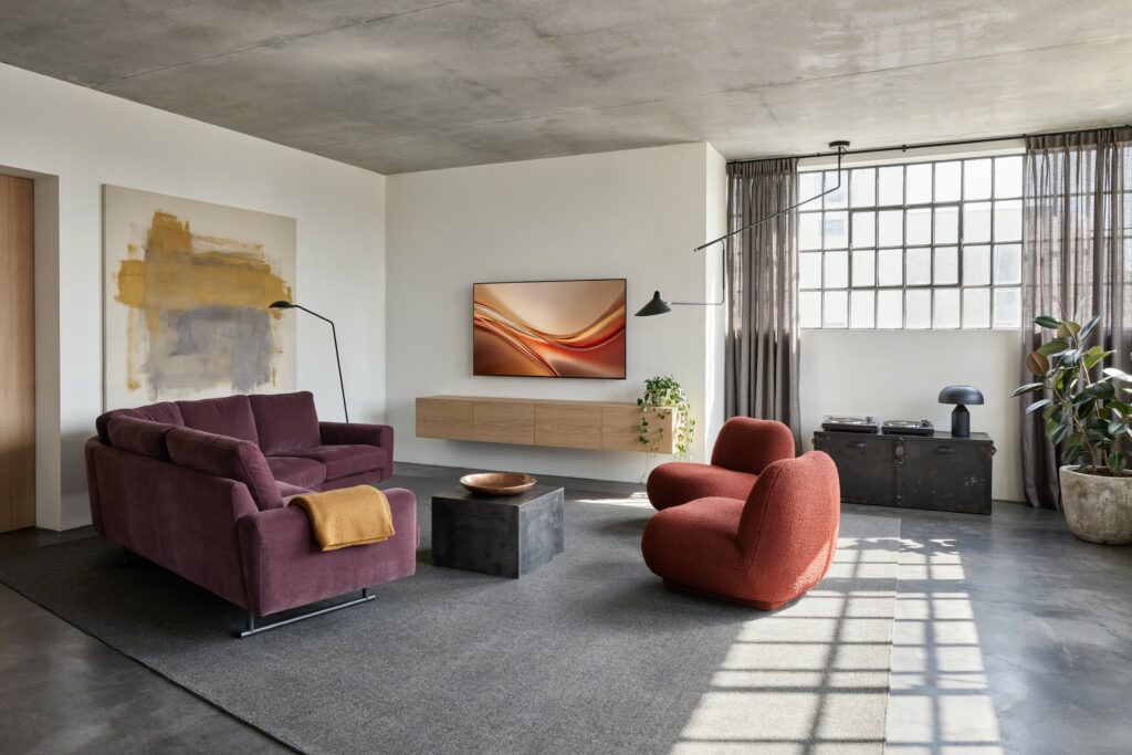 industrial living room with white-painted walls, a plum L-shaped sofa, rust-red lounge chairs, concrete ceiling and flooring, a gray rug, and a light wood floating media console