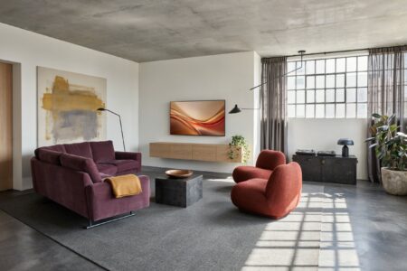 industrial living room with white-painted walls, a plum L-shaped sofa, rust-red lounge chairs, concrete ceiling and flooring, a gray rug, and a light wood floating media console