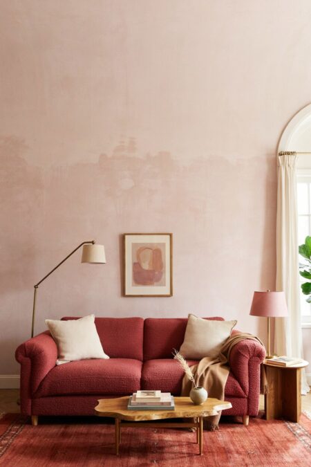 living room with pale pink wall behind the red sofa, a brick-red rug; cream throw pillows, a lamp shade, and curtains