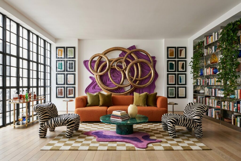 Modern maximalist living room with a burnt orange boucle sofa, zebra chairs, oversize 3D wall decor, and layered rugs. Vertical columns of small abstract art prints in glossy black frames. Stainless steel built-in shelving unit.