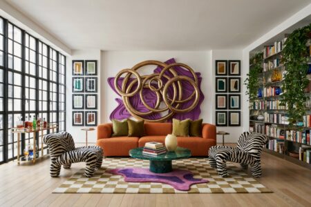 Modern maximalist living room with a burnt orange boucle sofa, zebra chairs, oversize 3D wall decor, and layered rugs. Vertical columns of small abstract art prints in glossy black frames. Stainless steel built-in shelving unit.