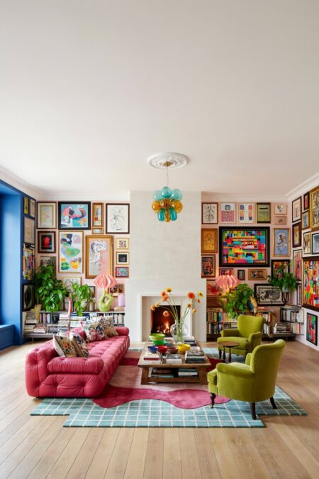 Modern maximalist living room with colorful glass pendant, a pink tufted sofa, green accent chairs, a gallery wall, and a fireplace. There is a blue accent wall on the side of the window.