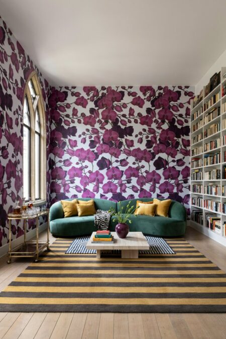 modern maximalist living room with purple floral wallpaper, striped rugs, a green velvet sofa with yellow pillow and a zebra print throw, and white bookshelves