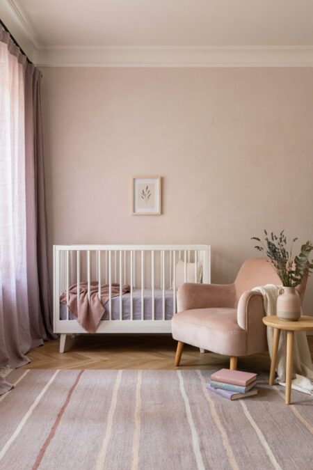 nursery with light pink walls, a pink velvet armchair, a pale lavender crib sheet, pale lavender curtains, and a rug