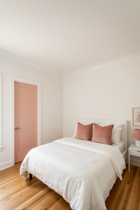 small bedroom with white walls and bedding; blush pink pillows and pink-painted door